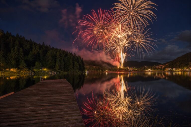 Feu d'artifice aux éclats dorés et cramoisis illuminant le ciel nocturne au-dessus d'un lac de montagne bordé de pins, avec un ponton en bois au premier plan.