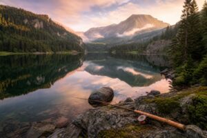 Lac de montagne paisible entouré de sapins et de pics brumeux dans les Vosges à l'aube, avec une canne à pêche posée sur la rive rocheuse au premier plan et la lumière dorée se reflétant sur l'eau calme.