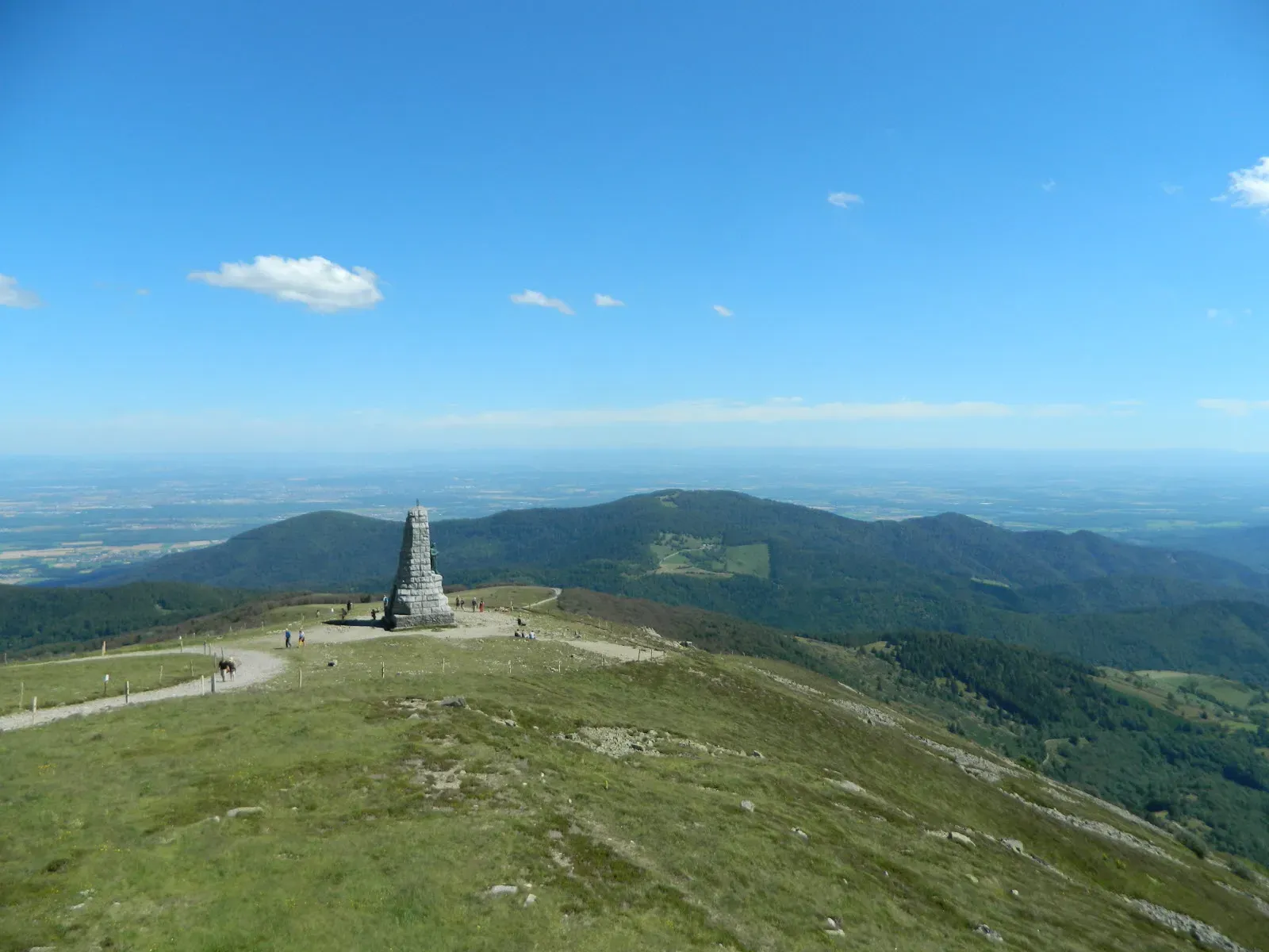 Route de montagne sinueuse sur une crête élevée avec de vastes vallées verdoyantes, des nuages printaniers, des sommets herbeux parsemés de fleurs sauvages, un cycliste solitaire au loin, et en bas un texte superposé