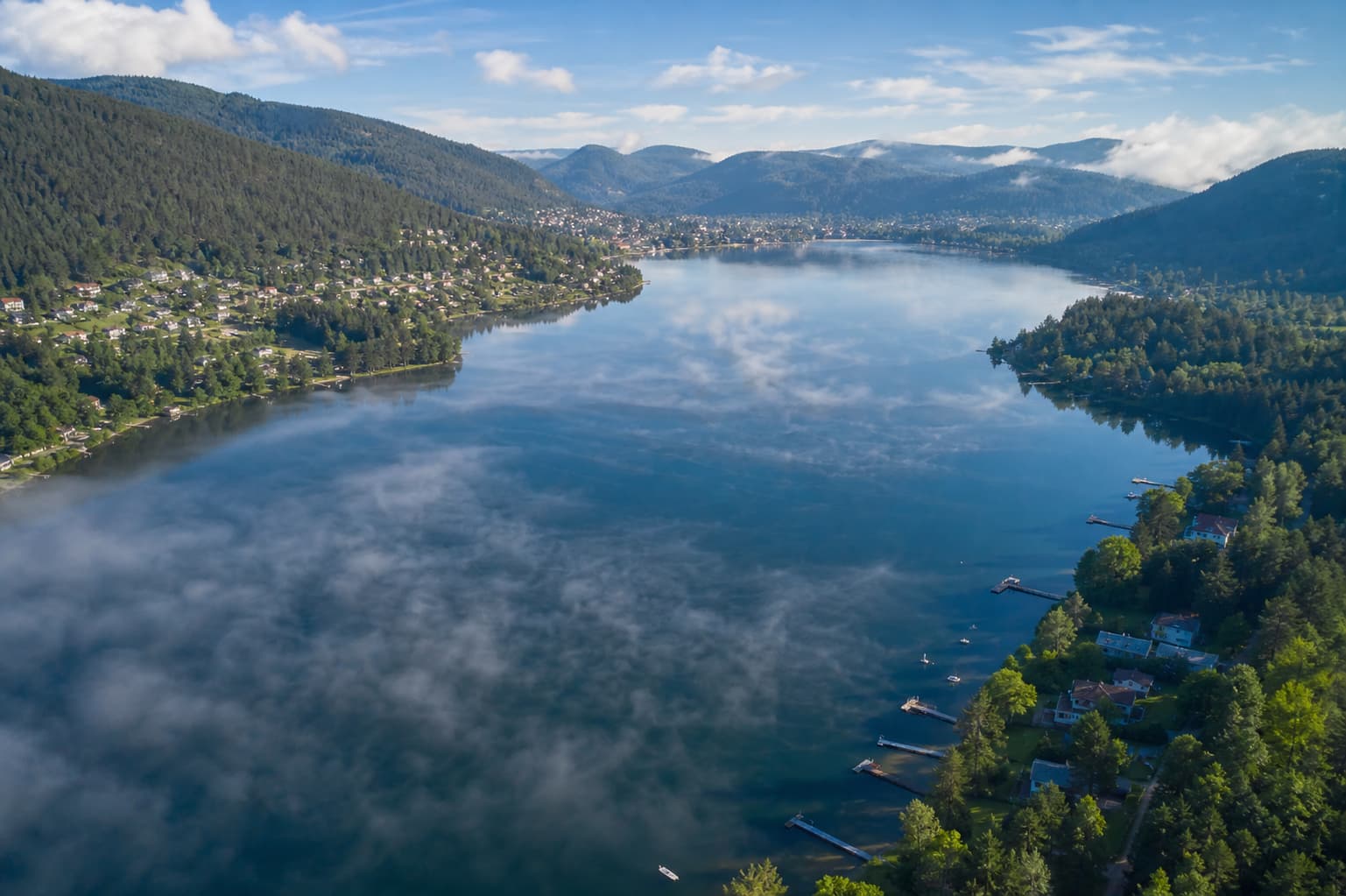 Vue aérienne du lac de Gérardmer, eau bleu-vert reflétant le ciel, rives boisées avec pontons et plates-formes de pêche, montagnes en arrière-plan et brume matinale.