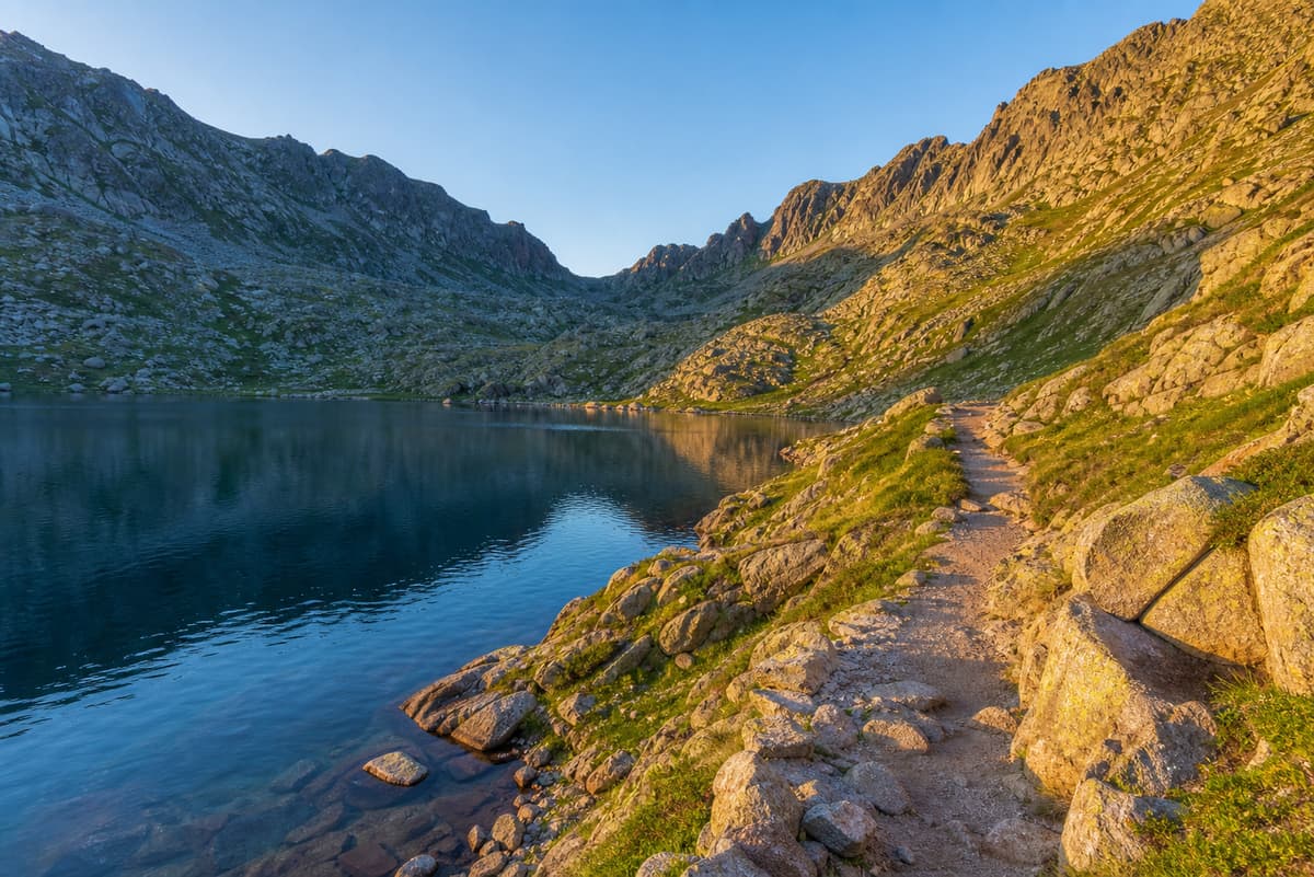 Lac de montagne aux rives rocheuses, entouré de pentes raides, avec un sentier visible sous une lumière de fin d’après-midi.